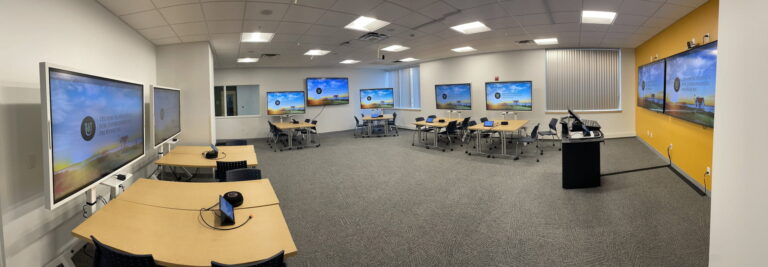 Wide view of a training room with displays at every table cluster and additional screens along both walls.