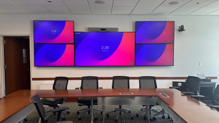 Executive board room with a five-screen display array, conferencing camera, and microphones set into the table.