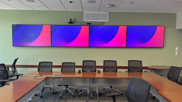 Board room with a four-display video wall, ceiling microphones, and a conferencing camera above the center displays.